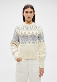 Jovem mulher com cabelo escuro e liso, vestindo um suéter creme com padrões geométricos azuis e bege, combinado com calças brancas, contra um fundo liso.