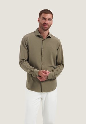 Man draagt een olijfgroen, lange mouw, knoophemd en witte broek, hij verstelt zijn linker manchet met een neutrale achtergrond.