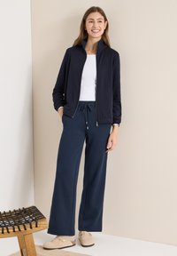 Granatowa kurtka na zamek na białym topie, zestawiona z granatowymi spodniami o szerokich nogawkach. Jasne buty wsuwane, drewniane krzesło na pierwszym planie.