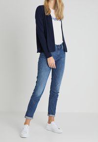Marinblå öppen cardigan över en vit t-shirt med blå text, kombinerad med uppvikta slim-fit blåjeans och vita sneakers.