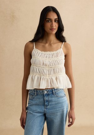 Mujer con cabello largo y negro, vestida con un top blanco sin mangas con volantes y detalles de encaje, y jeans azules de tiro alto, de pie frente a un fondo beige.