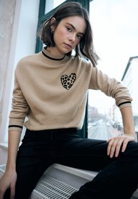 Maglione beige con un design a cuore con stampa leopardata, scollo a costine e dettagli a strisce nere. Indossato con jeans scuri, seduto contro una finestra.