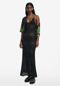 Black knitted maxi dress with a geometric pattern and a fitted design, paired with a floral short-sleeve shirt and platform sandals.