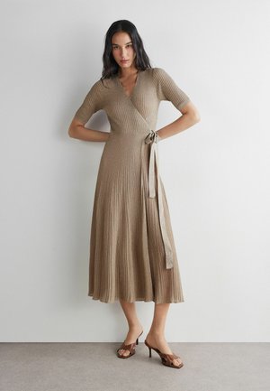 Frau mit langen dunklen Haaren, die ein beiges, geripptes Wickelkleid mit kurzen Ärmeln und braune Sandaletten mit Absatz trägt, steht vor einer schlichten Wand.