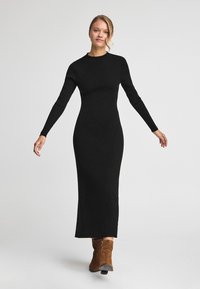 Svart ribbat stickat långärmat klänning med en åtsittande silhuett som sträcker sig ner till anklarna, tillsammans med bruna mockaankelboots.