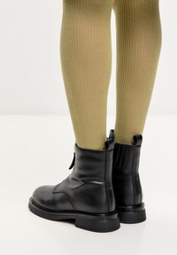 Stivaletti neri in pelle con una finitura liscia, pannelli laterali elasticizzati e suola piatta, indossati con leggings verde oliva a coste.