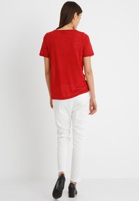 Röd t-shirt med kort ärm och rund halsringning, kombinerad med vita tajta byxor. Modellen har på sig svarta slip-on-skor. Mjuk, lätt tyg.