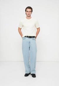 Camiseta blanca de manga corta, vaqueros de pierna ancha color azul claro, cinturón negro y zapatos negros. Fondo liso. Modelo de pie con las manos en los bolsillos.