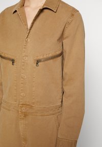 Gros plan sur une personne portant une combinaison beige à boutons, avec des poches poitrine zippées et un col, visible du cou à la taille.