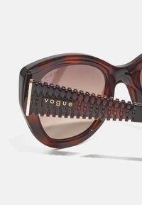 Tortoiseshell sunglasses with oversized cat-eye shape, gradient brown lenses, and textured black temples featuring a gold "vogue" logo.