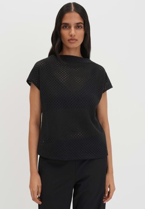 Donna con lunghi capelli scuri che indossa una maglia nera traforata a maniche corte e pantaloni neri, in piedi su uno sfondo chiaro.