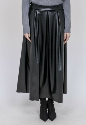 Personne portant une longue jupe en cuir noir plissée, un pull gris et des bottines noires, debout devant un fond clair uni.