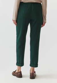 Green wool blend pants with a tapered shape, featuring rolled cuffs and back pockets. Styled with a light brown long-sleeve top and shoes.