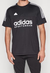 Černé sportovní tričko s bílým logem "adidas SPORTSWEAR". Má krátké rukávy, kulatý výstřih a horizontální žebrovanou strukturu se bočními pruhy.