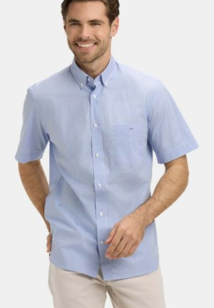Homme souriant portant une chemise à carreaux bleu clair à manches courtes avec une poche poitrine et un pantalon beige, debout avec les mains près des poches.