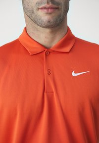 Oransje polo skjorte med klassisk krage, korte ermer og to-knapps placket. Har en hvit Nike-logo på venstre side.