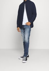 Tmavě modrá košile na knoflíky přes bílé tričko, doplněná světlými modrými džíny a černými teniskami s bílými podrážkami a tkaničkami. Neformální, vrstvený outfit.