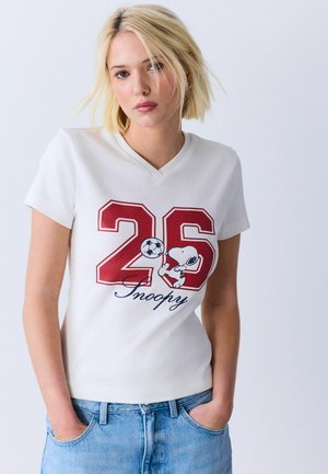Junge Frau mit kurzen blonden Haaren, die ein weißes T-Shirt mit roter Nummer 26 und Snoopy, der einen Fußball hält, trägt, blaue Jeans, Hände in den Taschen.