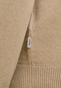 Gros plan d'un pull en tricot beige montrant une étiquette en tissu avec le texte "JJ REBEL" et un ourlet côtelé en bas.