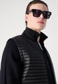 Μαύρο γιλέκο από πούπουλο με ραβδωτή υφή, σε συνδυασμό με μία μαύρη στρώση ψηλού γιακά. Μεγάλα, τετράγωνα μαύρα γυαλιά ηλίου προσθέτουν αντίθεση.