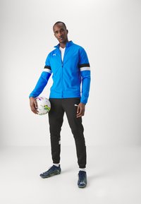Blauwe sweatshirt met rits en zwart-witte strepen, zwarte broek met witte accenten en blauwe voetbalschoenen. Houdt een kleurrijke voetbal vast.