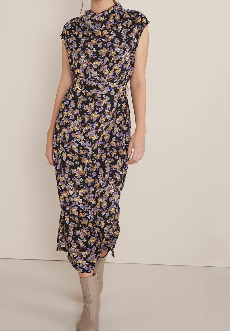Femme portant une robe midi noire à fleurs violettes et jaunes, taille ceinturée et bottines beige sur un fond uni.