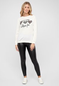 Krém színű pulóver, amelyen a "MINDNYÁJAN FÉNYLÜNK" szöveg látható grafikai díszítéssel és leopardmintás szívek találhatók, fekete leggings-szel és fehér sportcipővel párosítva.