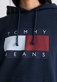 Felpa blu navy con un logo con la scritta "TOMMY" in bianco sopra una scatola azzurra e rossa. Ha un cappuccio con lacci e una consistenza morbida.