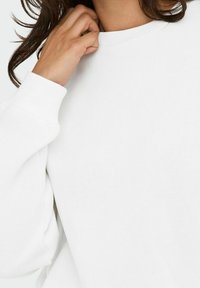 Felpa bianca con collo rotondo, polsini a costine e una texture liscia. Il tessuto appare morbido e informale, con dettagli di vestibilità comoda.