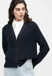 Γυναίκα με σγουρά σκούρα μαλλιά, ντυμένη με μπλε σκούρη ζακέτα με κουμπιά πάνω από άσπρο πουκάμισο με γιακά και ανοιχτό μπλε τζιν, που κοιτάζει προς τα πλάγια.