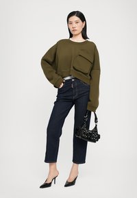 Sweatshirt scurt de culoare verde măslin, cu un buzunar frontal, asortat cu blugi de culoare albastră închisă și pantofi cu toc negri, ținând o geantă neagră cu accente.