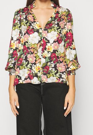 Kvinde iført en blomsterprintet bluse med lange ærmer i pink, hvid, gul og rød kombineret med sorte bukser med hvid stikning.