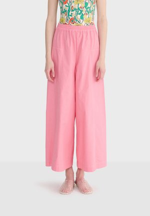 Personne portant un pantalon rose ample taille haute, un haut fleuri sans manches et des chaussures translucides tissées sans lacets, debout devant un fond uni.