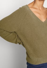 Olivgrüner, lockerer Pullover mit V-Ausschnitt, strukturiertem Strick und geripptem Saum. Verfügt über überschnittene Schultern und weite Ärmel.