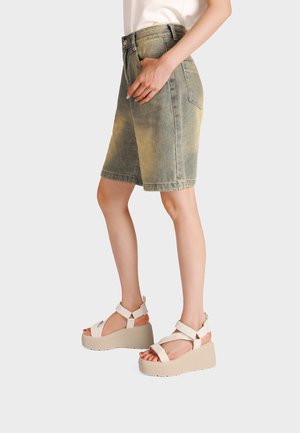 Modella che indossa pantaloncini di jeans verdi scoloriti fino al ginocchio e sandali platform beige con cinturini alla caviglia, in piedi davanti a uno sfondo semplice.