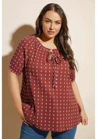 Blusa color ruggine con maniche corte, dettaglio con nodo al collo, tessuto testurizzato e piccoli pattern geometrici bianchi su tutta la superficie. Vestibilità casual e comoda.