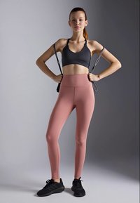 Ung kvinne i svart treningsbh og rosa leggings poserer selvsikkert mens hun holder et strikk mot en nøytral grå bakgrunn.