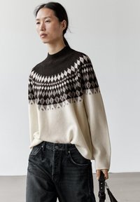 Femme portant un pull crème et marron à motifs avec un jean noir, tenant une sangle de sac noire dans un cadre intérieur minimaliste.
