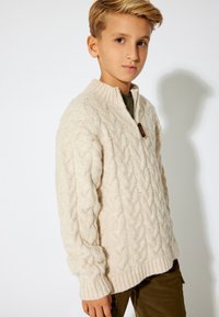 Cremefarbenes Strickpullover mit Kabelmuster, hohem Kragen und Viertel-Reißverschluss. Gerippte Bündchen und Saum sorgen für Struktur.