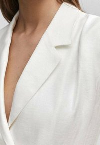 Biały lniany żakiet z głębokim dekoltem w kształcie litery V i wyraźnie zarysowanymi ramionami. Posiada gładką fakturę oraz czysty, minimalistyczny design.