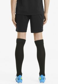 Sorte atletiske shorts parret med knæ høje sorte sokker og blå fodboldstøvler med grønne accenter på en ensfarvet baggrund.