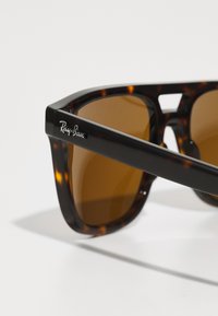 Ochelari de soare tortoiseshell maro cu lentile dreptunghiulare. Au un cadru negru, cu un branding proeminent pe tâmple și o textură netedă.