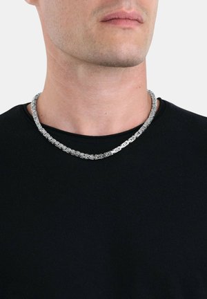 Silberne Kettenhalskette mit ineinandergreifenden Gliedern, getragen über einem schwarzen T-Shirt. Merkmale eine glatte, glänzende Textur und ein einheitliches Design.