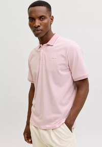 Mann trägt ein hellrosa kurzärmeliges Poloshirt und cremefarbene Hose, steht mit einer Hand in der Tasche vor einfachem Hintergrund.