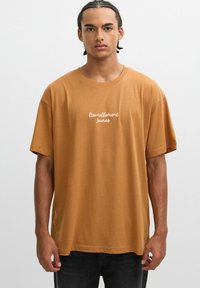 Camiseta de algodón marrón de manga corta, con 'Éternellement Jeunes' impreso en letras cursivas blancas en el pecho. Textura suave.
