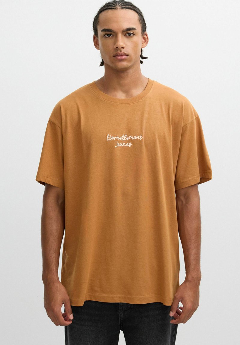Camiseta de algodón marrón de manga corta, con 'Éternellement Jeunes' impreso en letras cursivas blancas en el pecho. Textura suave.
