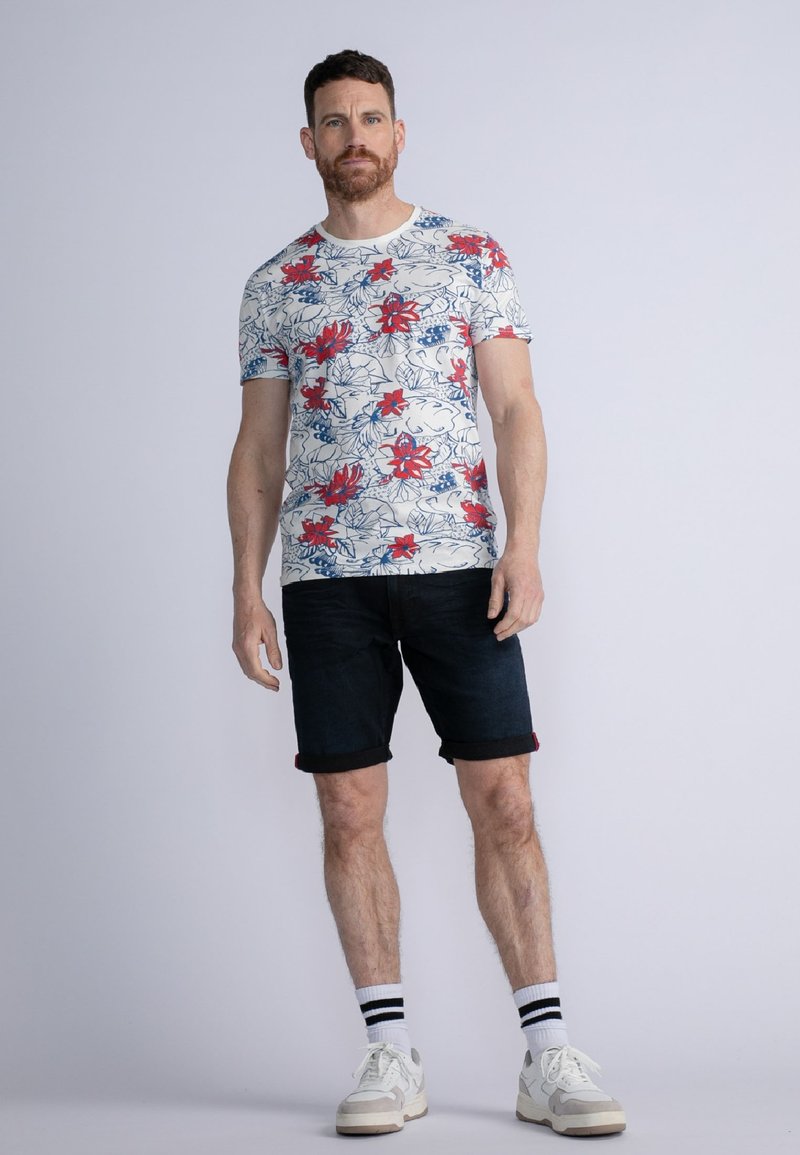 Camiseta de manga corta con cuello redondo que presenta un estampado floral con flores rojas sobre un fondo blanco. Combinada con pantalones cortos de mezclilla oscuros y remangados.