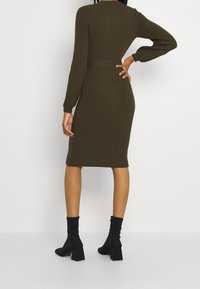 Kvinna som bär en ribbad mörkgrön midi-sweaterklänning och svarta klackiga ankelstövlar, poserar med en hand på höfterna, sedd bakifrån.