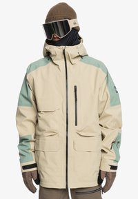 Giacca da sci beige con accenti turchesi, materiali impermeabili, cappuccio regolabile, numerose tasche e cerniera nera, indossata con un berretto marrone e occhiali da sci.