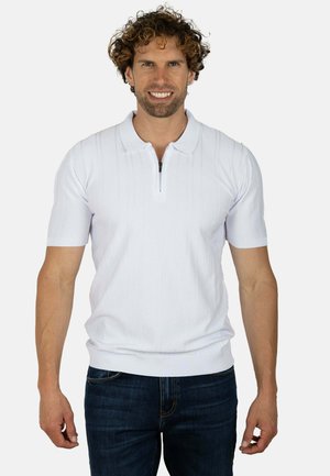 Hombre con el pelo rizado sonríe, lleva un polo blanco de manga corta con cremallera y vaqueros oscuros, de pie frente a un fondo claro y liso.